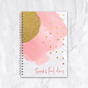 May include: A spiral-bound notebook with a pink and gold abstract design. The cover features a pink watercolour wash, gold glitter accents, and gold polka dots. The text "Sarah's food diary" is written in pink script.
