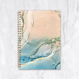 May include: A spiral-bound notebook with a watercolour design. The cover features a blend of peach, blue, and gold hues, creating an abstract, flowing pattern. The spiral binding is gold-coloured.