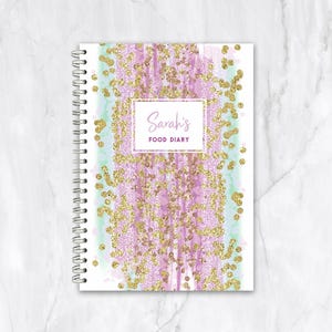 May include: A spiral-bound food diary with a decorative cover. The cover features a watercolour design in shades of purple and mint green, accented with gold glitter. The text "Sarah's FOOD DIARY" is displayed in a rectangular frame.