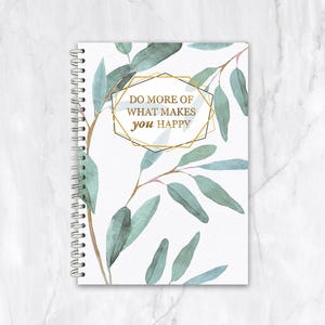 May include: A spiral-bound notebook with a white cover. It features watercolour eucalyptus leaves and the text "DO MORE OF WHAT MAKES you HAPPY" in a gold geometric frame. The leaves are green and blue.
