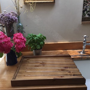 May include: A wooden draining board with a grooved surface, sitting on a wooden countertop next to a white porcelain sink. The countertop is made of reclaimed wood and has a rustic look. A silver faucet is visible in the background.