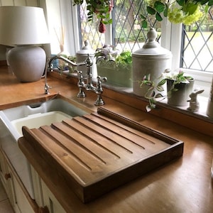 May include: A wooden draining board with grooves for drying dishes, placed next to a white porcelain kitchen sink. The sink is in a kitchen with a wooden countertop and a white cabinet.