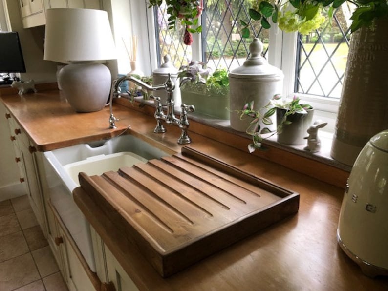 May include: A wooden draining board with grooves for drying dishes, placed next to a white porcelain kitchen sink. The sink is in a kitchen with a wooden countertop and a white cabinet.