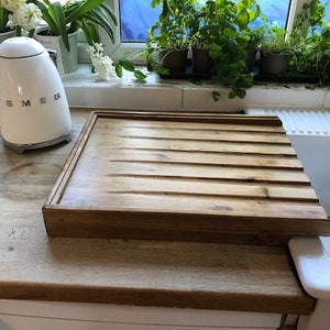 May include: A wooden dish drying rack with a grooved surface sits on a countertop next to a white ceramic sink. The rack is made of natural wood and has a rustic look.