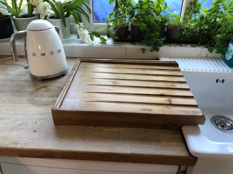 May include: A wooden dish drying rack with a grooved surface sits on a countertop next to a white ceramic sink. The rack is made of natural wood and has a rustic look.