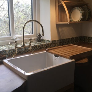 May include: A white porcelain kitchen sink with a gold-toned faucet and a wooden cutting board. The sink is set against a backdrop of colorful patterned tiles.