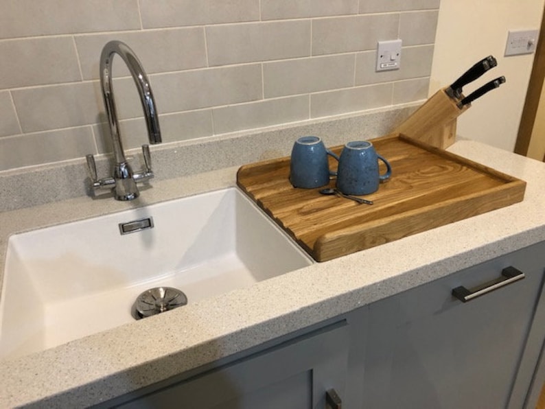 May include: A white kitchen sink with a chrome faucet and a wooden cutting board with two blue mugs on top. The cutting board is sitting on a gray countertop.