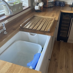 May include: A white porcelain farmhouse sink with a wooden draining board. The sink has a blue dishcloth inside. The draining board is made of wood and has a slatted design.