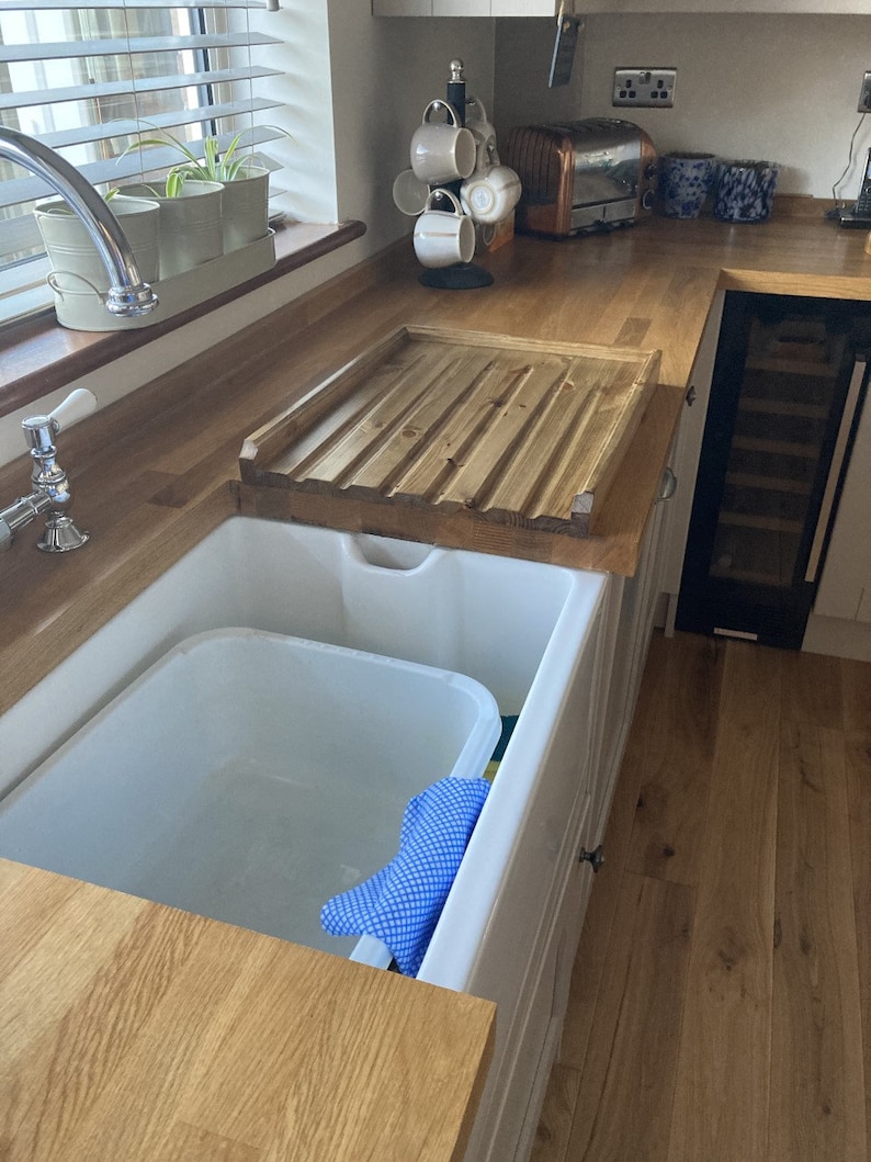 May include: A white porcelain farmhouse sink with a wooden draining board. The sink has a blue dishcloth inside. The draining board is made of wood and has a slatted design.
