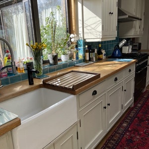 May include: A white farmhouse style kitchen sink with a wooden draining board and white cabinets. The sink is set against a backdrop of a window with a view of a garden.