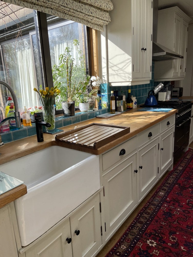 May include: A white farmhouse style kitchen sink with a wooden draining board and white cabinets. The sink is set against a backdrop of a window with a view of a garden.