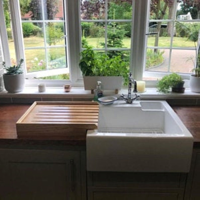 May include: A white farmhouse sink with a wooden cutting board that slides over the sink. The sink is in a kitchen with a window behind it.