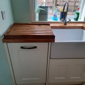 May include: A white kitchen sink with a wooden cutting board on the counter above it. The sink is made of porcelain and has a single bowl. The cutting board is made of wood and has a grooved surface. There is a white cabinet below the sink and a white cabinet to the left of the sink.
