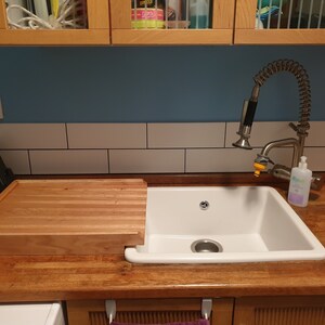 May include: A white porcelain kitchen sink with a wooden cutting board that sits on the counter next to the sink. The sink has a single drain and a chrome faucet with a pull-out sprayer.