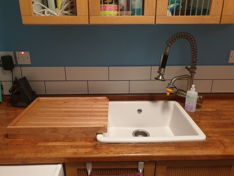 May include: A white porcelain kitchen sink with a wooden cutting board that sits on the counter next to the sink. The sink has a single drain and a chrome faucet with a pull-out sprayer.