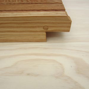 May include: A close-up of a wooden cutting board with a natural finish. The board has a rectangular shape with rounded edges and a groove running along the length of the board. The board is made of light-colored wood with a grain pattern.