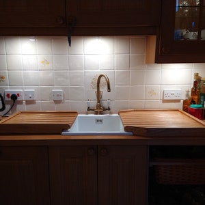 May include: A white porcelain kitchen sink with a brass faucet and two wooden draining boards. The sink is set against a white tiled backsplash with a decorative pattern.