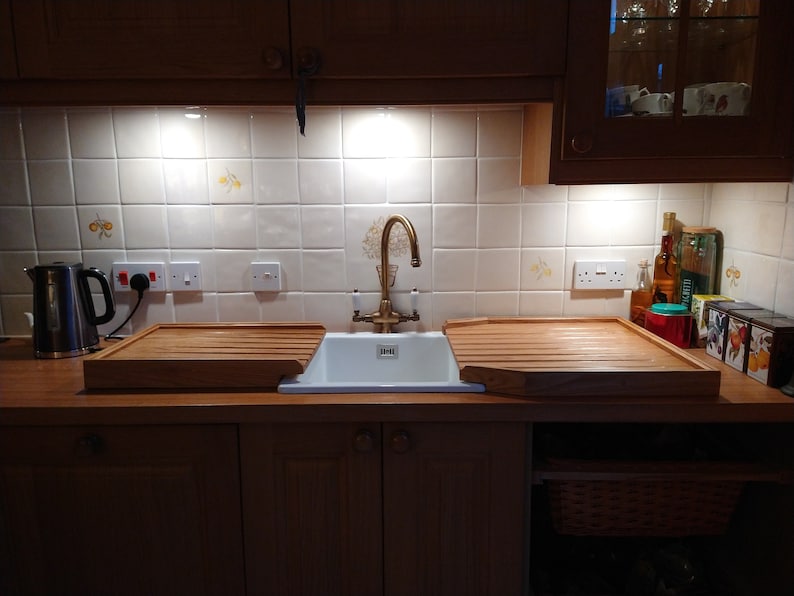 May include: A white porcelain kitchen sink with a brass faucet and two wooden draining boards. The sink is set against a white tiled backsplash with a decorative pattern.