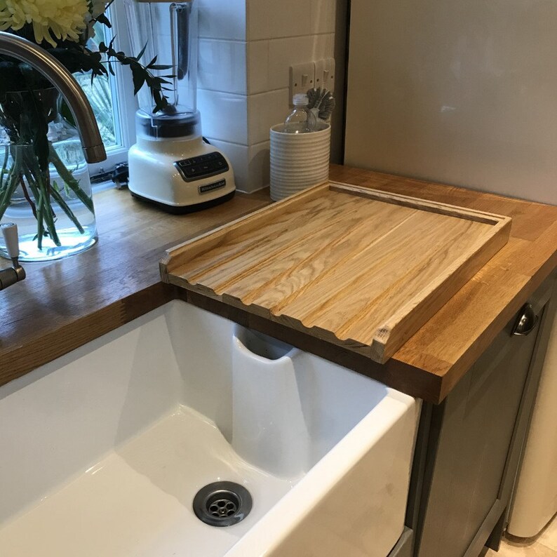 May include: A wooden cutting board with a groove on the edge sits on a countertop next to a white porcelain sink. The cutting board is made of light wood with a natural grain pattern.