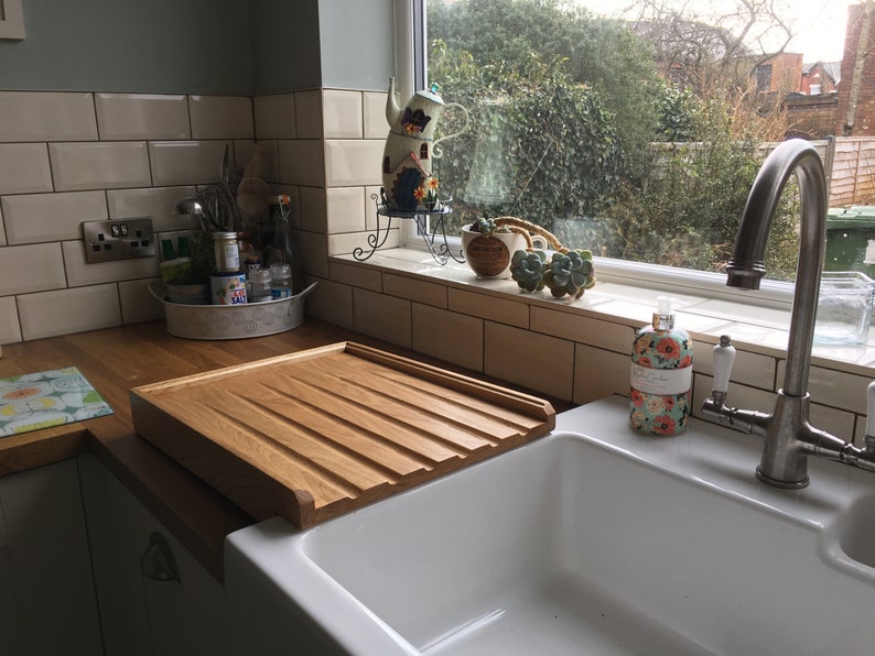 May include: A white porcelain kitchen sink with a wooden draining board. The sink has a chrome faucet and a bottle of soap with a floral design. The sink is set against a backdrop of white subway tiles.