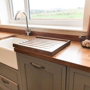 May include: A white porcelain farmhouse sink with a wooden draining board and a chrome faucet. The sink is set into a gray countertop with gray cabinets below.