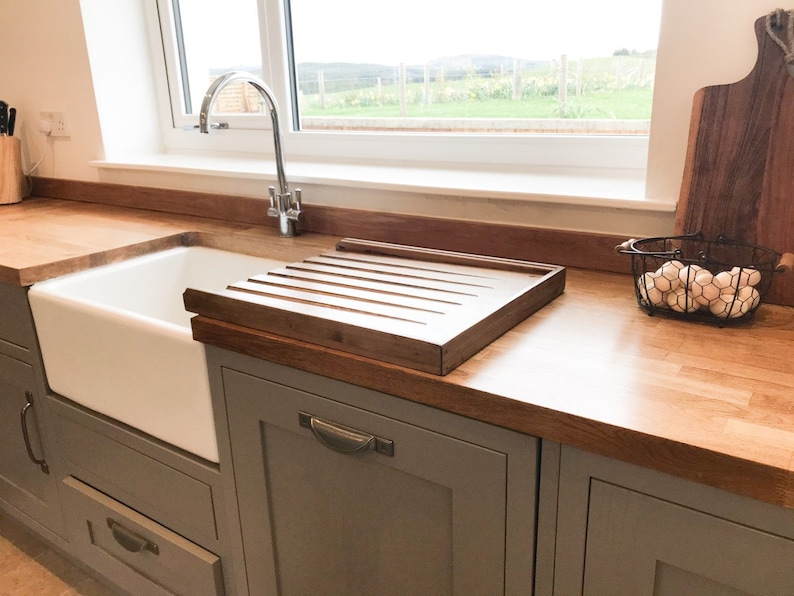 May include: A white porcelain farmhouse sink with a wooden draining board and a chrome faucet. The sink is set into a gray countertop with gray cabinets below.