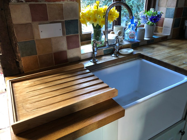 May include: A white porcelain farmhouse sink with a wooden draining board. The sink is set into a wooden countertop with a tiled backsplash. A bouquet of yellow daffodils is in a glass vase on the windowsill.