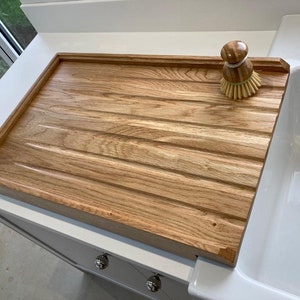 K&ouml;nnte beinhalten: Ein Holz-Abtropfgestell mit gerillter Oberfl&auml;che und erh&ouml;htem Rand. Das Gestell steht neben einem wei&szlig;en Porzellansp&uuml;lbecken. Eine gelbe und braune Sp&uuml;lb&uuml;rste liegt auf dem Gestell.
