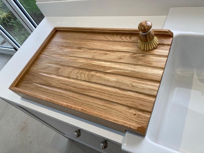 K&ouml;nnte beinhalten: Ein Holz-Abtropfgestell mit gerillter Oberfl&auml;che und erh&ouml;htem Rand. Das Gestell steht neben einem wei&szlig;en Porzellansp&uuml;lbecken. Eine gelbe und braune Sp&uuml;lb&uuml;rste liegt auf dem Gestell.