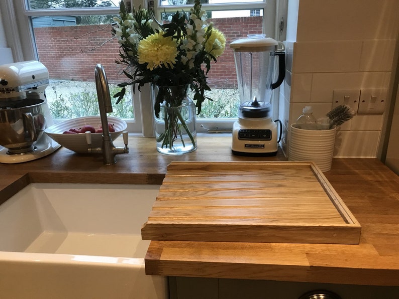 May include: A white kitchen sink with a wooden cutting board that slides over the sink. A white stand mixer and a white blender are on the counter next to a window.