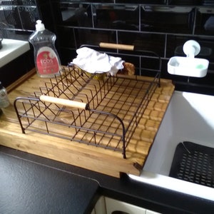 May include: A wooden dish drying rack with a metal wire insert and a wooden handle. The rack is sitting on a black countertop next to a white porcelain sink. There are two bottles of dish soap on the counter, one green and one red.