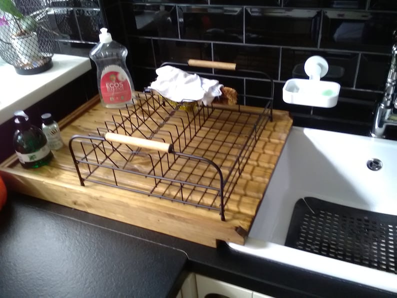 May include: A wooden dish drying rack with a metal wire insert and a wooden handle. The rack is sitting on a black countertop next to a white porcelain sink. There are two bottles of dish soap on the counter, one green and one red.