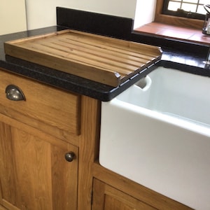 May include: A wooden cutting board with grooves sits on a black countertop next to a white porcelain farmhouse sink. The sink is part of a wooden cabinet with a drawer and a door.