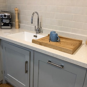 May include: A white kitchen sink with a gray cabinet underneath. The sink has a chrome faucet and a wooden cutting board with two blue mugs on it. There is a white tile backsplash and a knife block with black knives on the counter.