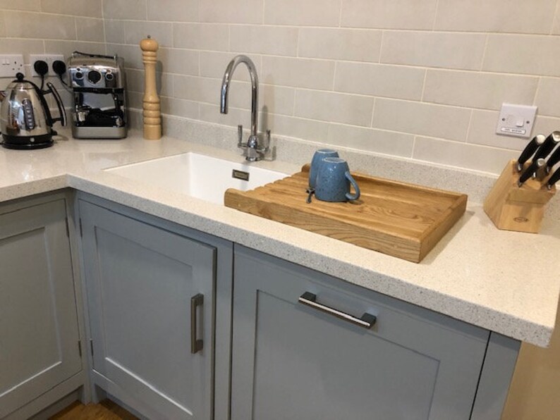 May include: A white kitchen sink with a gray cabinet underneath. The sink has a chrome faucet and a wooden cutting board with two blue mugs on it. There is a white tile backsplash and a knife block with black knives on the counter.