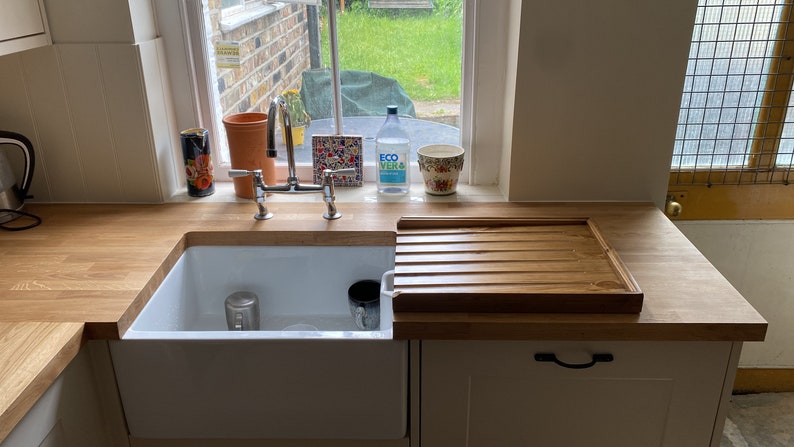 May include: A white porcelain kitchen sink with a wooden cutting board next to it. The sink is set into a wooden countertop. There are two chrome faucets and a window behind the sink.