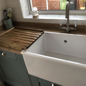 May include: A white ceramic farmhouse sink with a wooden draining board. The sink has a single bowl and a brushed nickel faucet with two handles. The draining board is made of wood and has slats to allow water to drain.