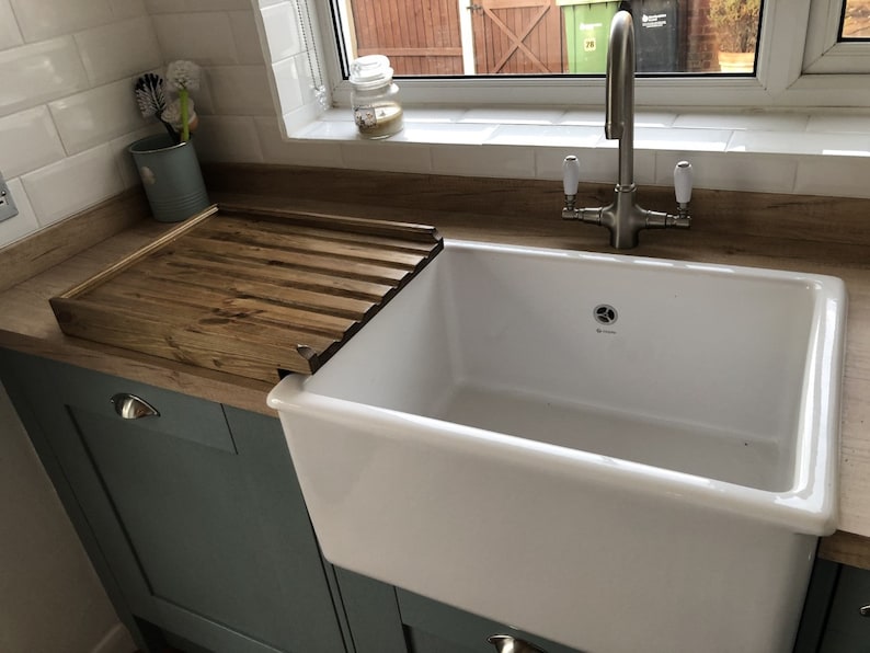May include: A white ceramic farmhouse sink with a wooden draining board. The sink has a single bowl and a brushed nickel faucet with two handles. The draining board is made of wood and has slats to allow water to drain.