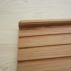 May include: A close-up of a light brown wood panel with a raised edge and horizontal grooves. The panel is made of natural wood with a smooth finish.