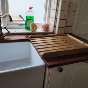 May include: A white porcelain farmhouse sink with a copper faucet and a wooden draining board. The sink is set into a countertop made of dark wood.