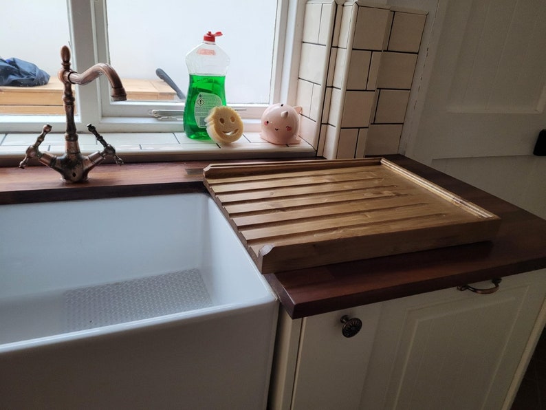 May include: A white porcelain farmhouse sink with a copper faucet and a wooden draining board. The sink is set into a countertop made of dark wood.
