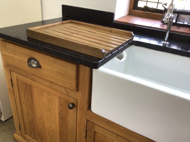 May include: A wooden cutting board with grooves sits on a black countertop next to a white farmhouse sink. The sink is made of porcelain and has a single bowl.