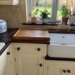 May include: A white ceramic bread box with the word "Bread" printed in blue on the front. The bread box is sitting on a black countertop next to a wooden cutting board. The cutting board is sitting on a white cabinet with brown knobs. A white porcelain sink with a gold faucet is in the background.