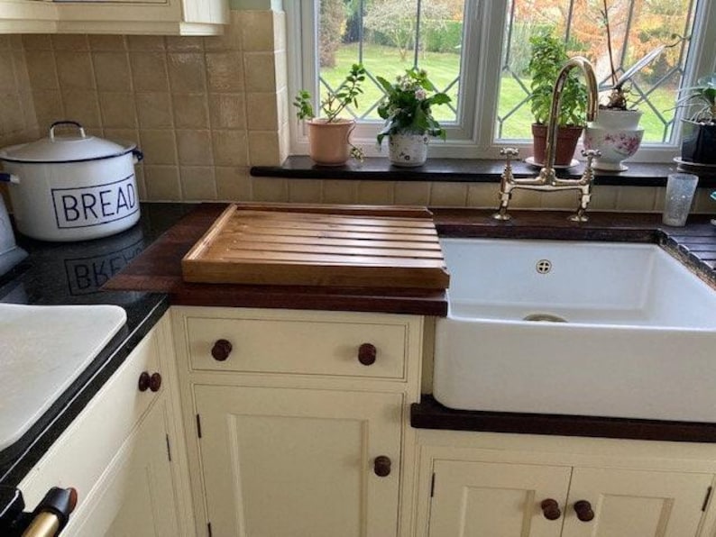 May include: A white ceramic bread box with the word "Bread" printed in blue on the front. The bread box is sitting on a black countertop next to a wooden cutting board. The cutting board is sitting on a white cabinet with brown knobs. A white porcelain sink with a gold faucet is in the background.
