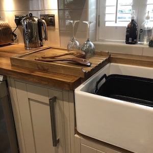 May include: A white farmhouse sink with a black plastic container inside. A wooden cutting board with two wooden spoons sits on the counter next to the sink. A silver electric kettle is on the counter behind the cutting board.