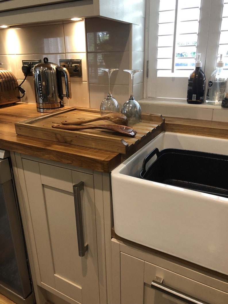 May include: A white farmhouse sink with a black plastic container inside. A wooden cutting board with two wooden spoons sits on the counter next to the sink. A silver electric kettle is on the counter behind the cutting board.