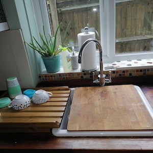 May include: A white kitchen sink with a wooden cutting board on top. There is a wooden dish rack with cups and mugs on the counter next to the sink. A plant is on the windowsill above the sink.