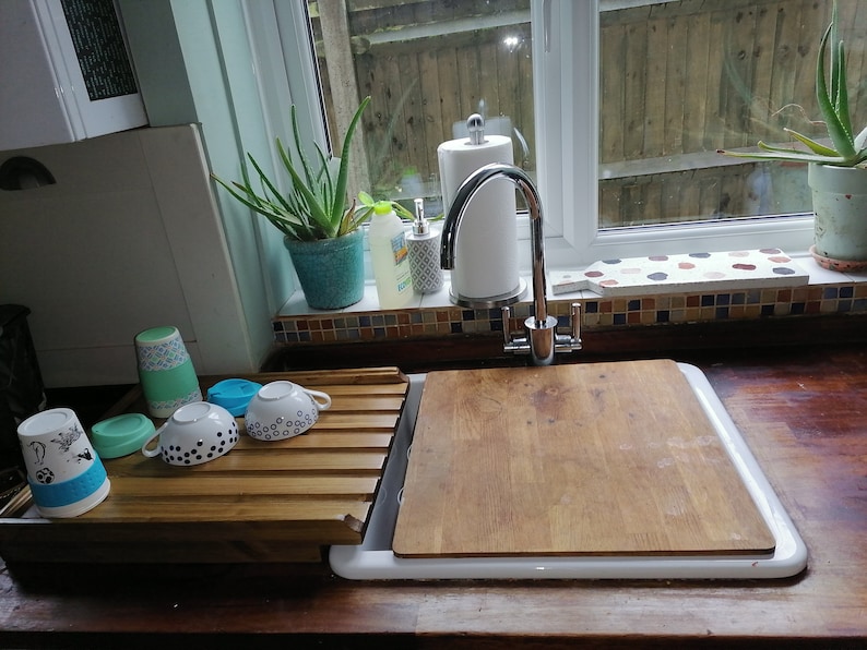 May include: A white kitchen sink with a wooden cutting board on top. There is a wooden dish rack with cups and mugs on the counter next to the sink. A plant is on the windowsill above the sink.