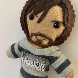 May include: A felt doll of a man with brown hair and a beard, wearing a gray and black striped shirt. The doll has a name tag that reads "Azkaban Prison XY390".