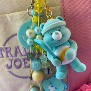 May include: A keychain featuring a light blue flocked bear figurine holding a tennis racket, accented with yellow star charms, a light blue smiley face disc, and a strand of iridescent, light blue, and yellow beads on a gold metal ring.